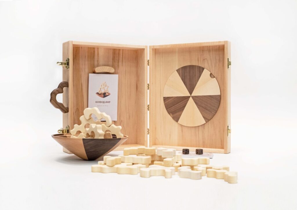 A wooden tabletop game set called Genderloop displayed open, showing a round board, wooden game pieces, dice, and an instruction booklet.
