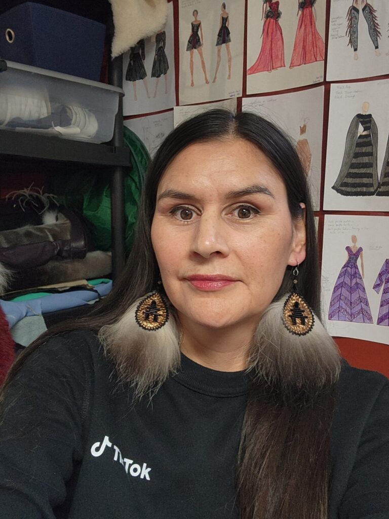A person with long dark hair wears beaded and fur earrings, standing in a studio space with fashion sketches pinned to the wall behind.