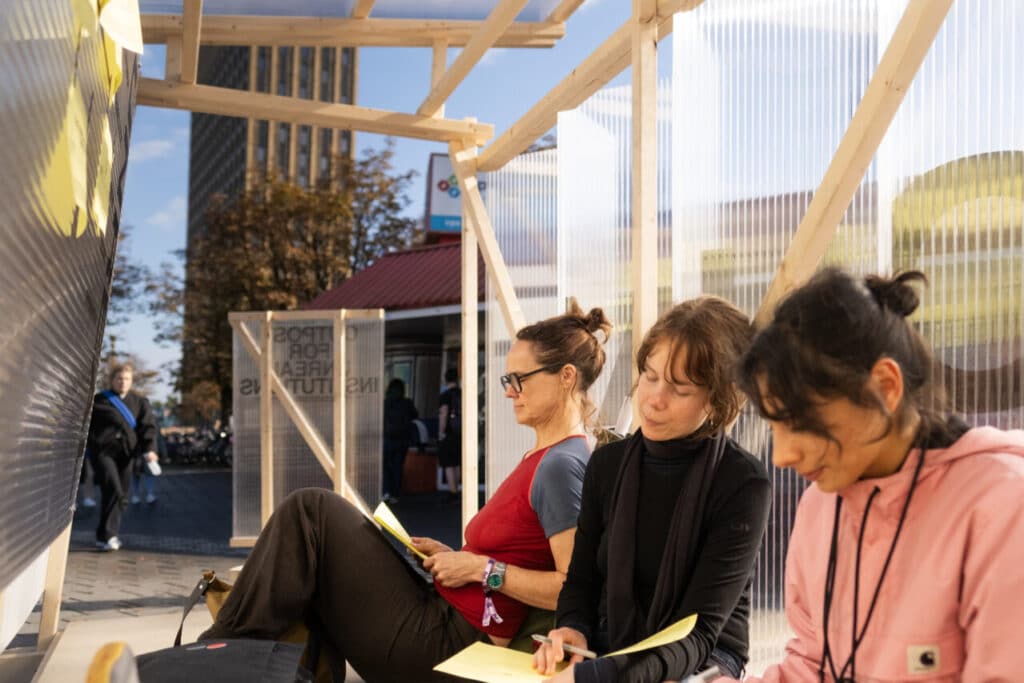 Three people sitting inside a wooden pavilion structure, writing on yellow sheets of paper during a public activity.