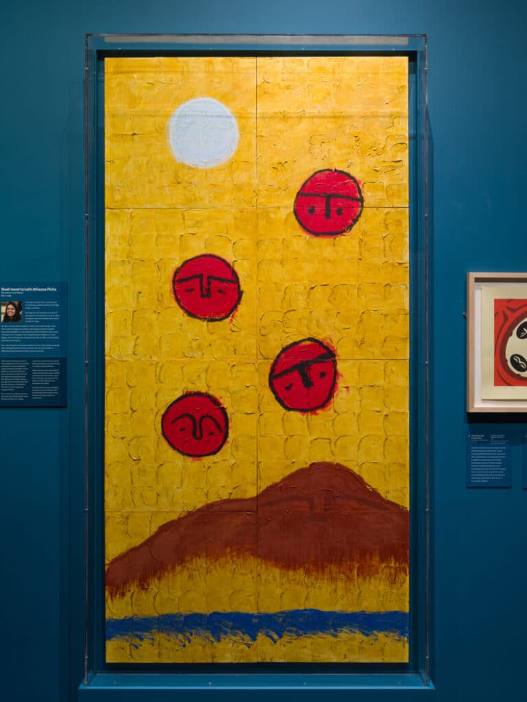 Large painting with yellow background, red faces, a white circle, brown hill and blue strip at the bottom.