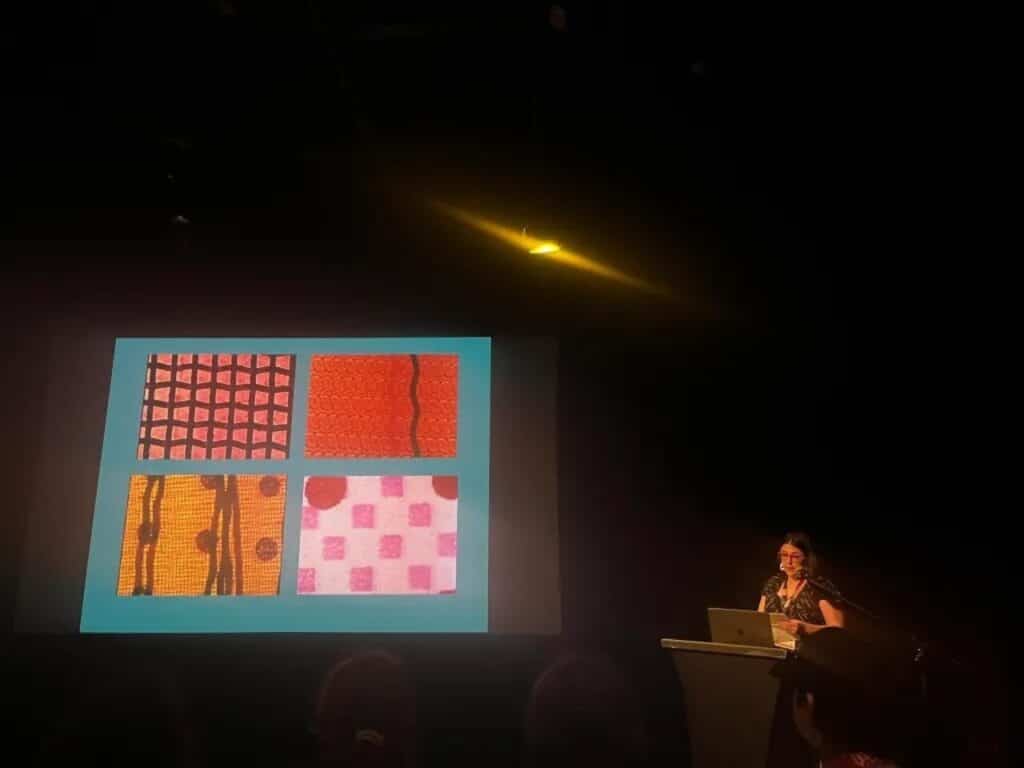 Person at a podium presenting slides with four colourful abstract patterns projected on a screen.