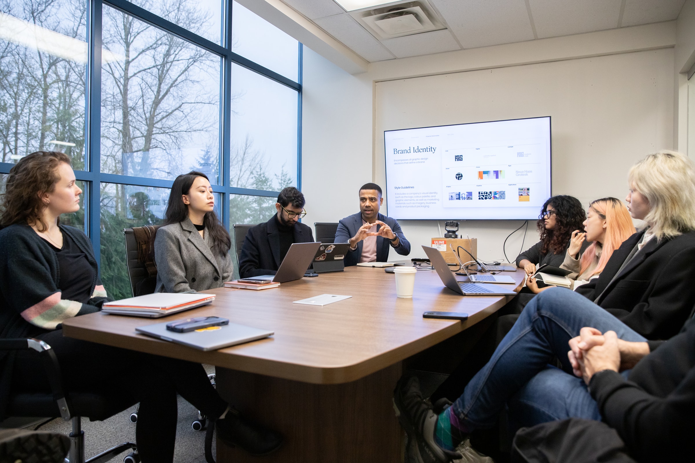 A group of people sit around a conference table in a meeting room, listening to a presenter. A large screen behind them displays a slide titled “Brand Identity.”