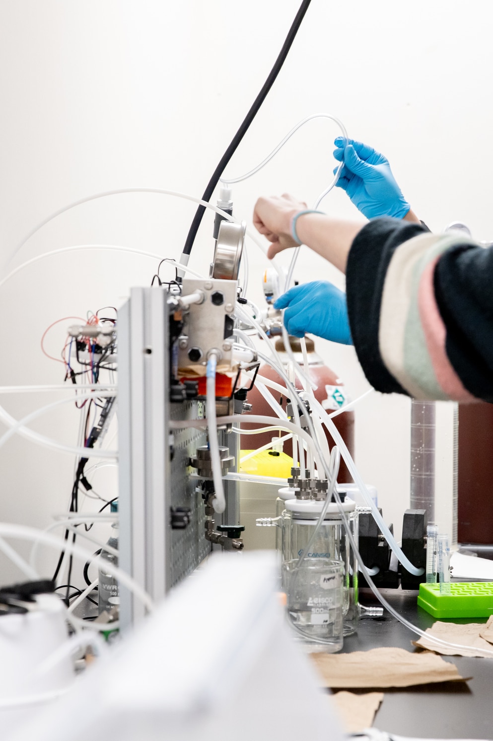 Gloved hands adjust tubes and glassware on a laboratory machine filled with wires, tubing, and beakers.