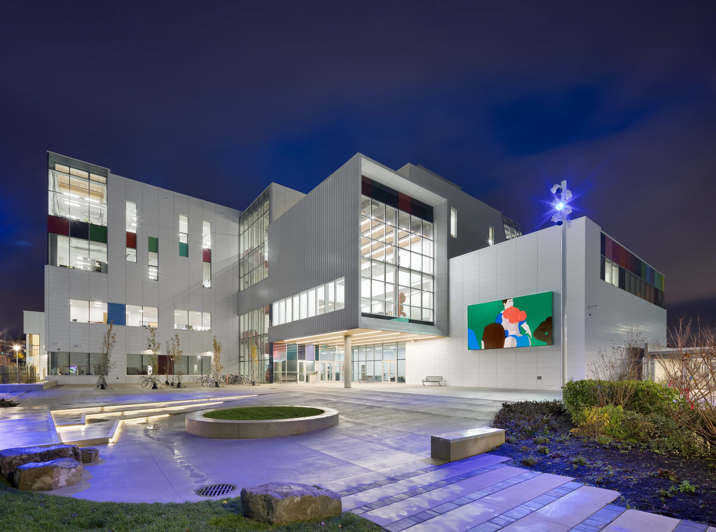 ECU exterior at night, lit by architectural lights, with illuminated digital art on the facade and landscaped seating in the courtyard.