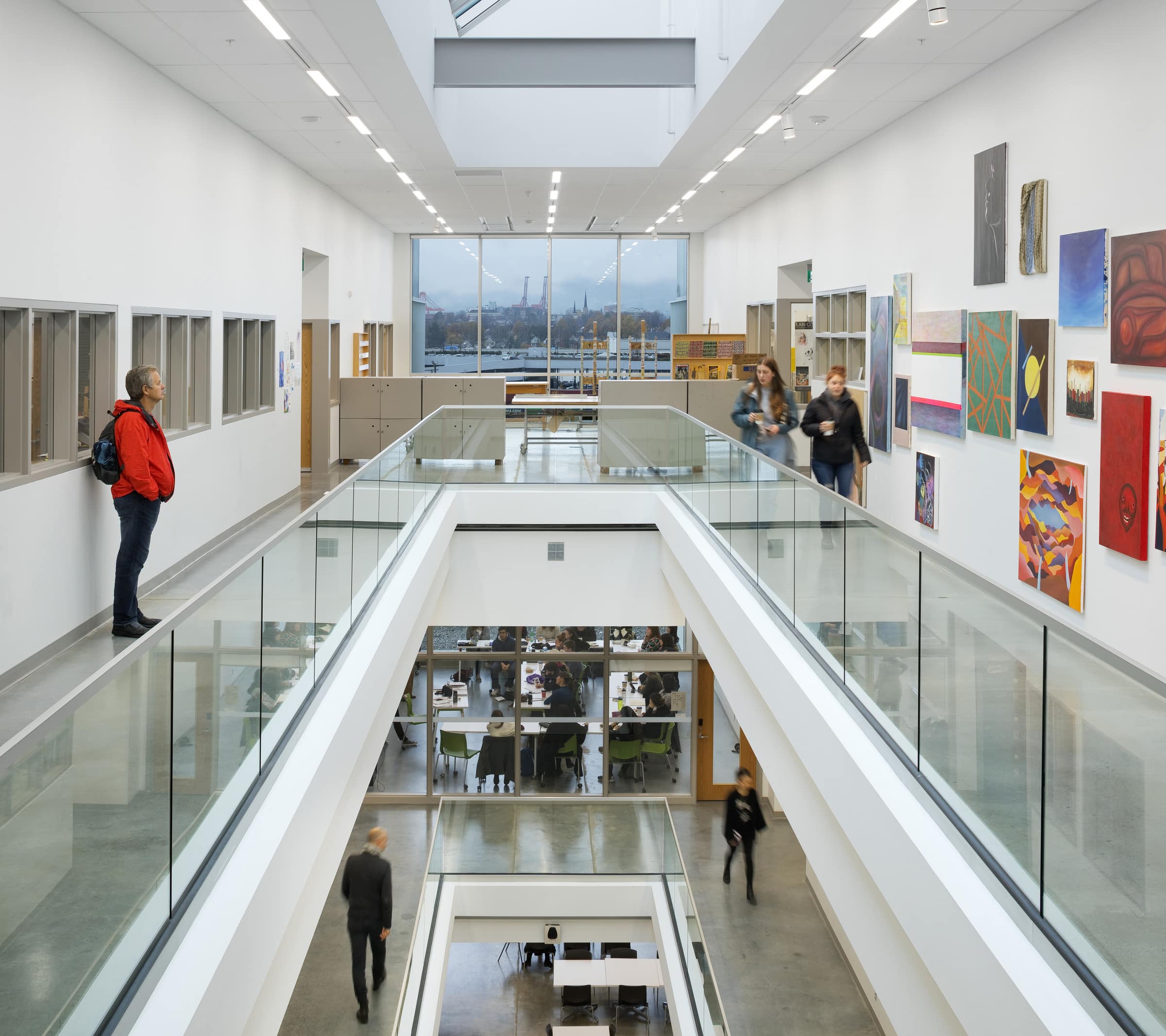 Modern lounge with black armchairs, carved wooden art, and large windows overlooking an urban view. A person reads on a couch while others work at tables in the background.