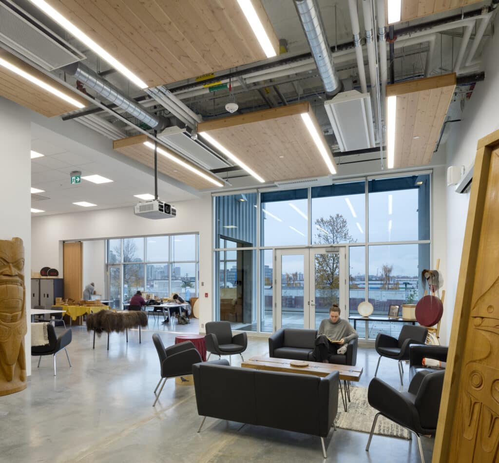 Modern university common area with high ceilings and exposed ductwork, furnished with black armchairs around low tables. A person sits reading on the right, while two people work at a large table covered with hides in the background. Floor-to-ceiling windows reveal an overcast cityscape outside. Carved wooden panels and Indigenous art pieces decorate the space.