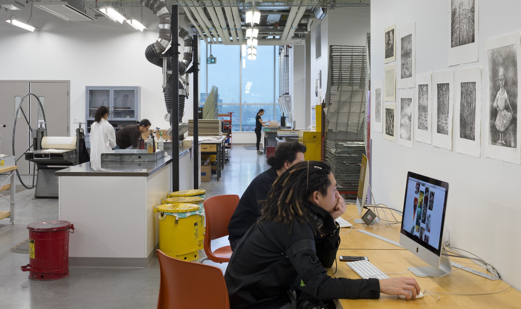 University printmaking studio with students working at computers and print stations. Artwork hangs on the walls, and large ventilation pipes run overhead.