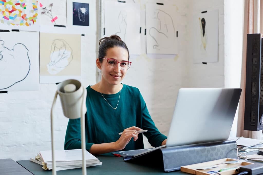 Designer at a desk with a laptop and digital pen, smiling in front of sketches and drawings pinned to the wall.