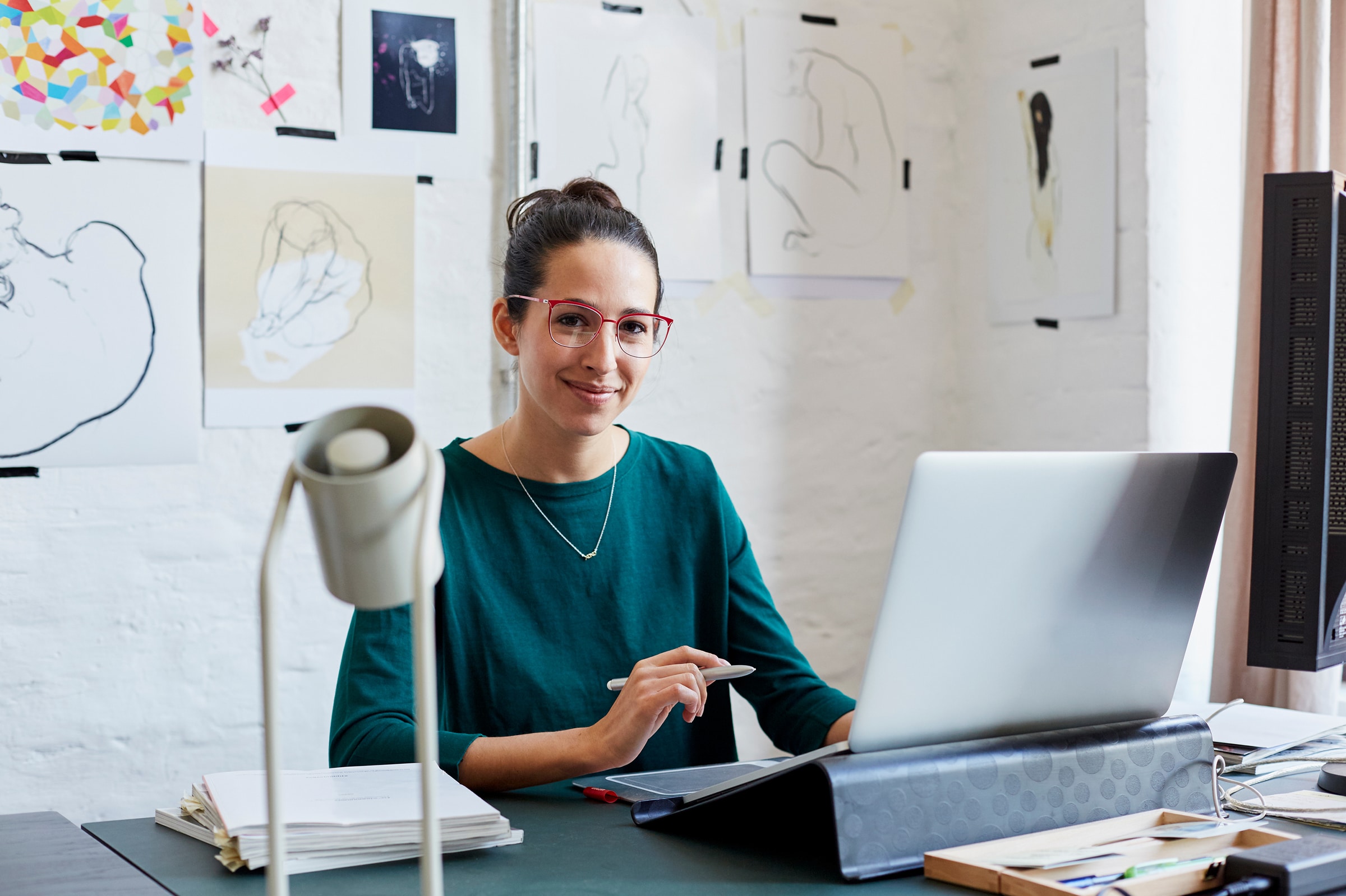 Designer at a desk with a laptop and digital pen, smiling in front of sketches and drawings pinned to the wall.