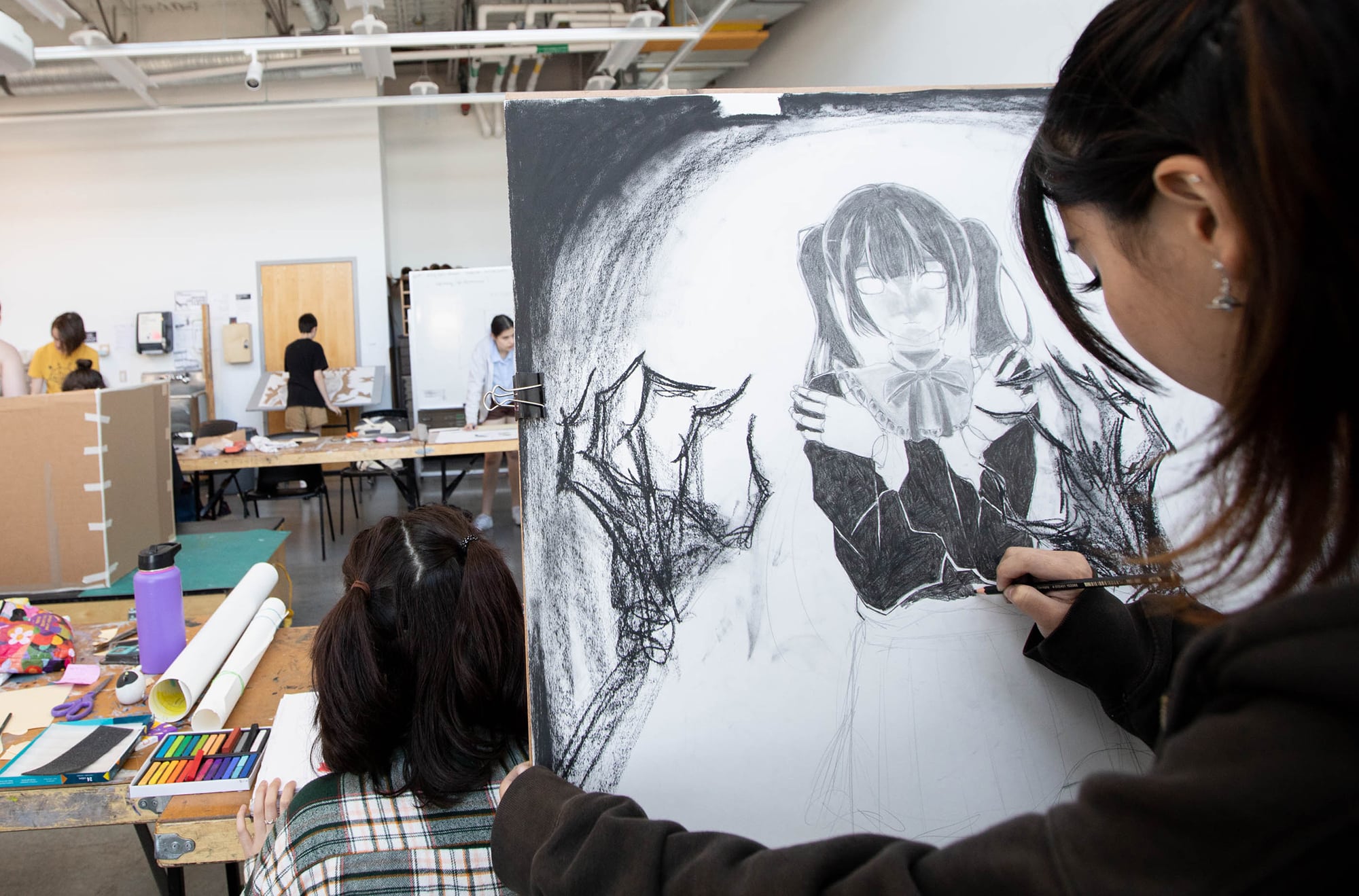 Student sketching dark fantasy figure — A student works on a large charcoal drawing of a gothic character with wings, seated in a busy studio.