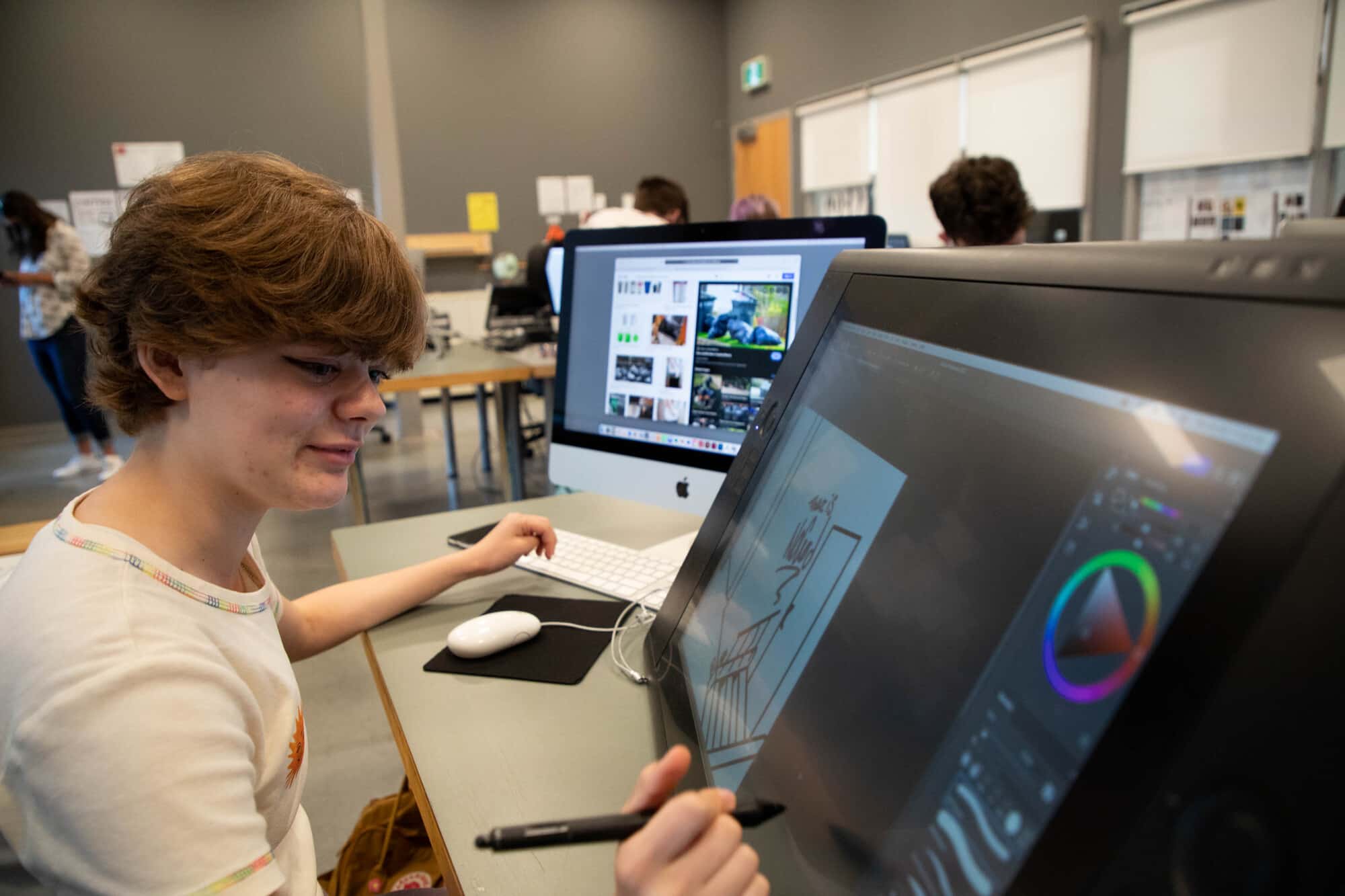 Student using digital drawing tablet — A teen uses a pen on a large drawing tablet while working on digital art, with a computer monitor open beside them.