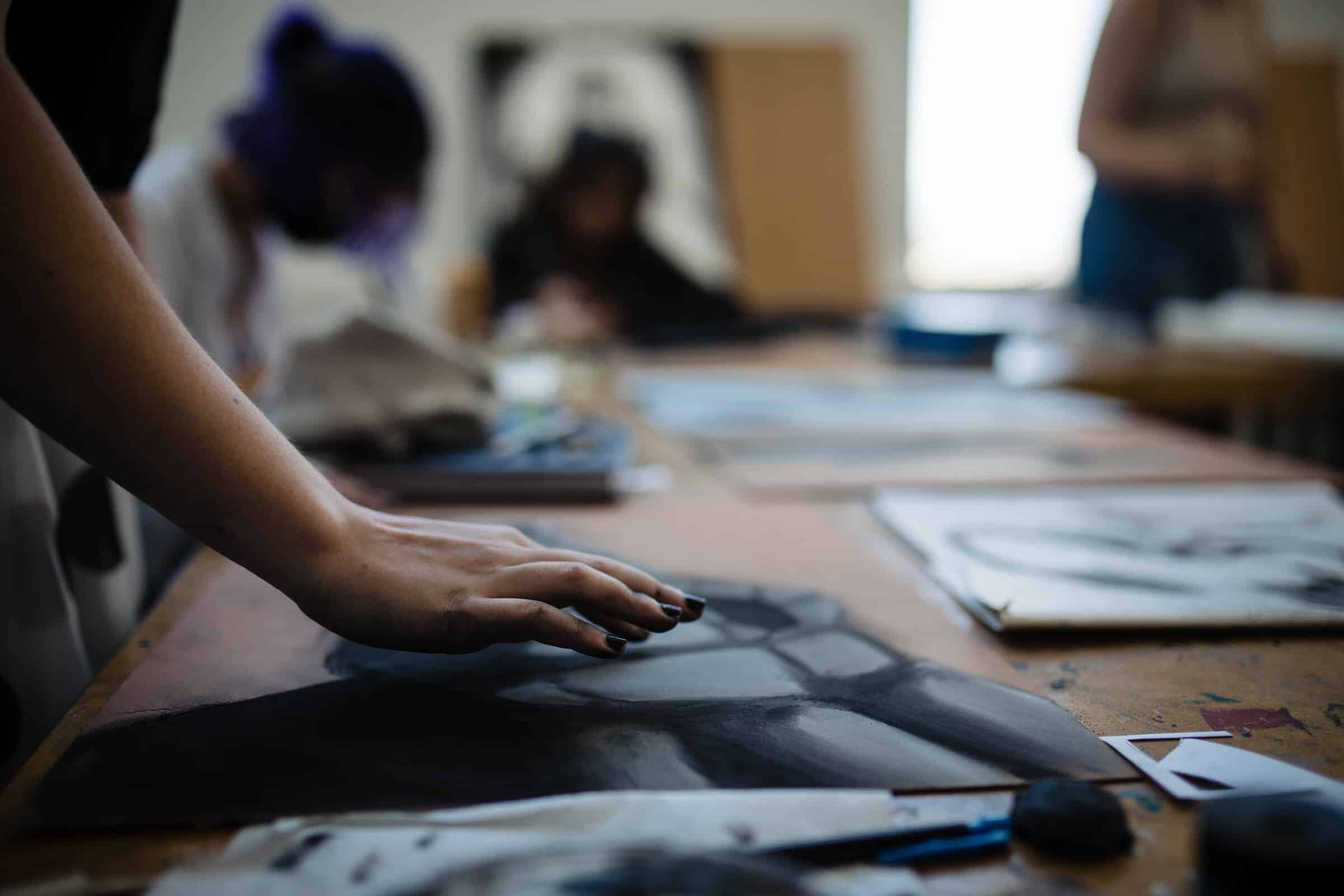Hand blending charcoal drawing — A hand presses and blends charcoal on a drawing of a geometric shape, with students sketching in the background.