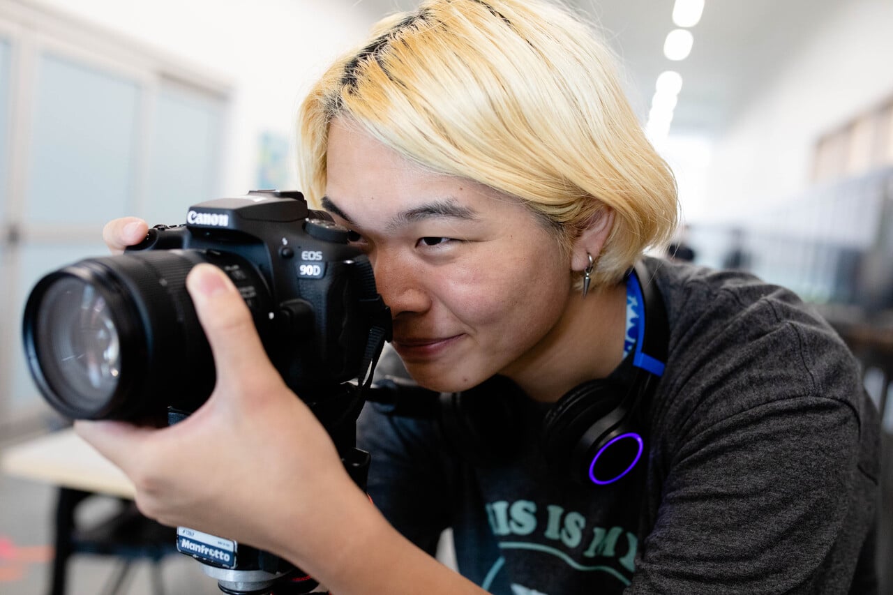 A student with blonde hair looking through the viewfinder of a Canon EOS 90D camera, preparing to take a photograph indoors.