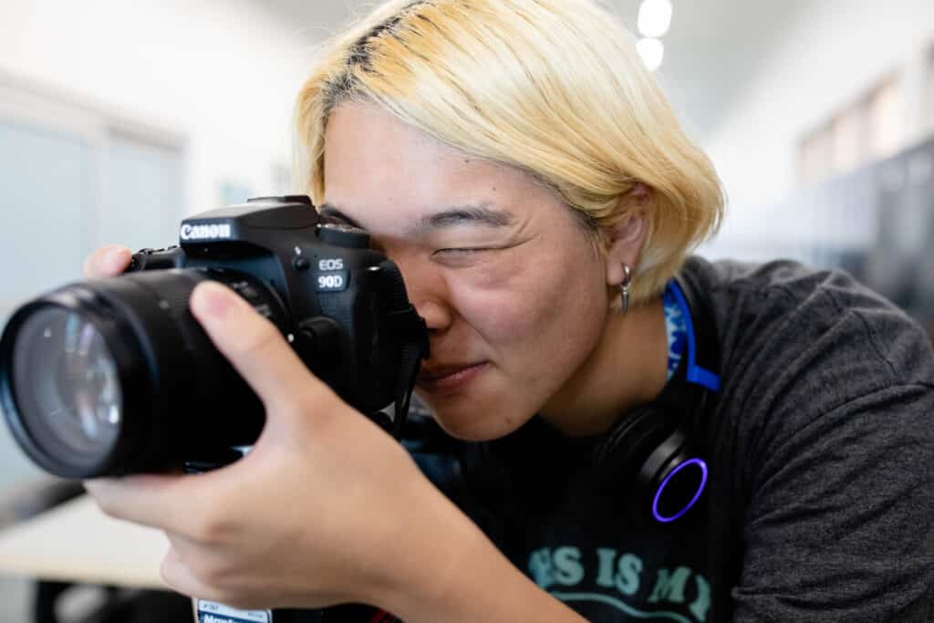 Close-up of a student with blond hair holding a Canon DSLR camera, looking through the viewfinder to capture a shot.