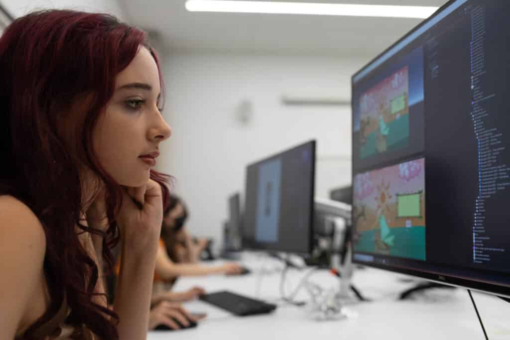 A student with long red hair looks intently at her computer screen, working on a project with colorful digital assets.