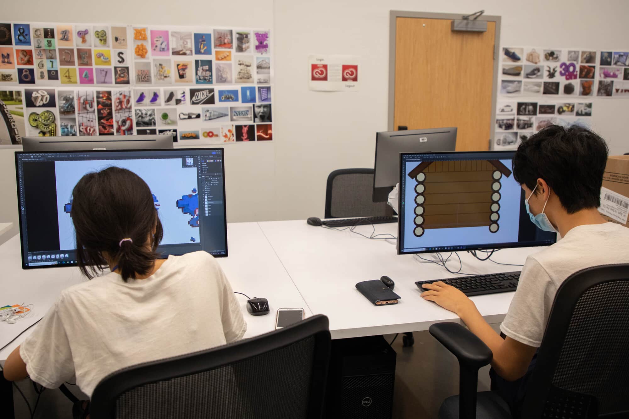Two students sit at desktop computers in a design lab, creating pixel art and vector graphics on large monitors.