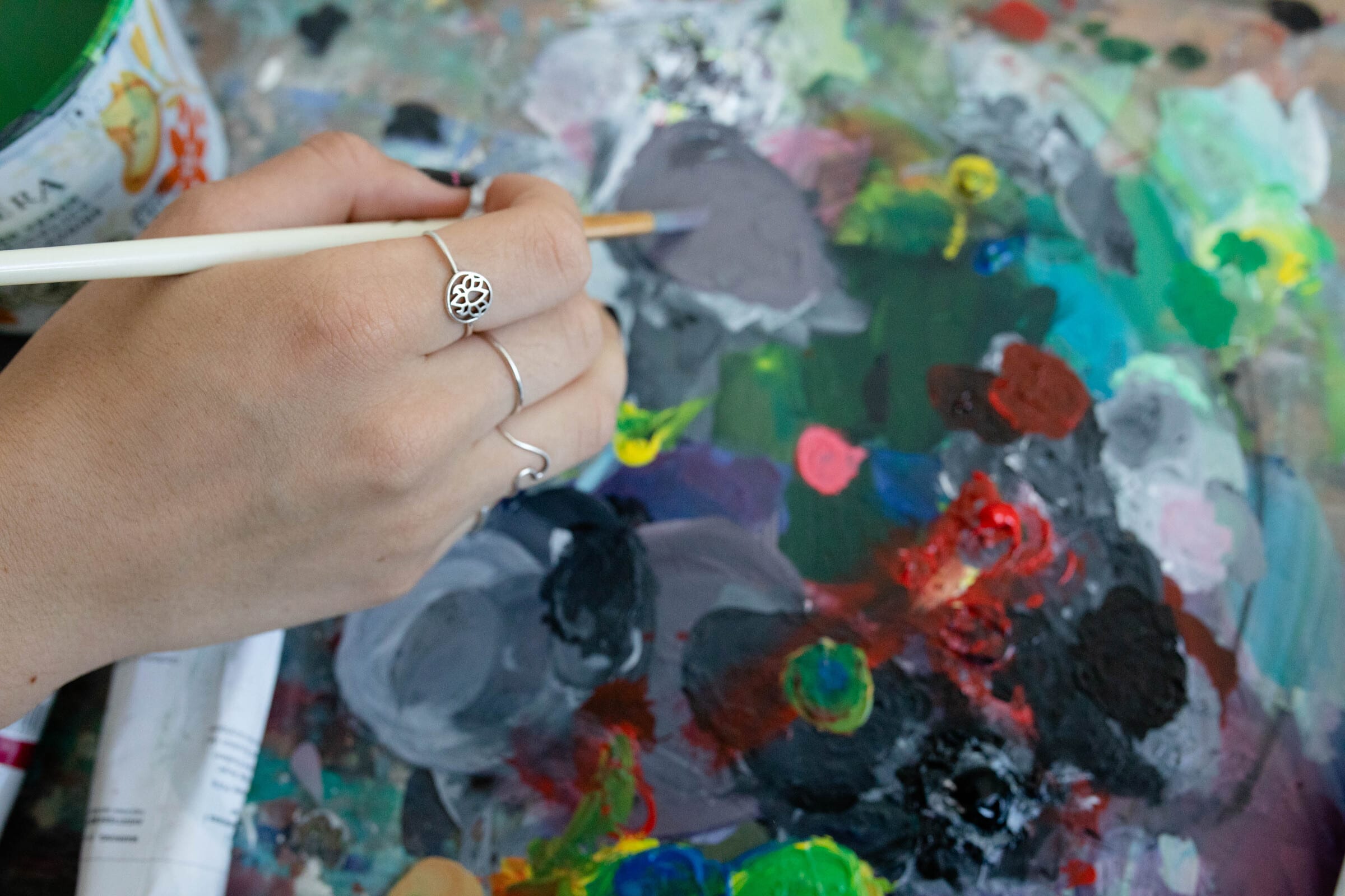 Close-up of a hand mixing paint on a messy, multicolored palette with a brush, surrounded by tubes of paint.