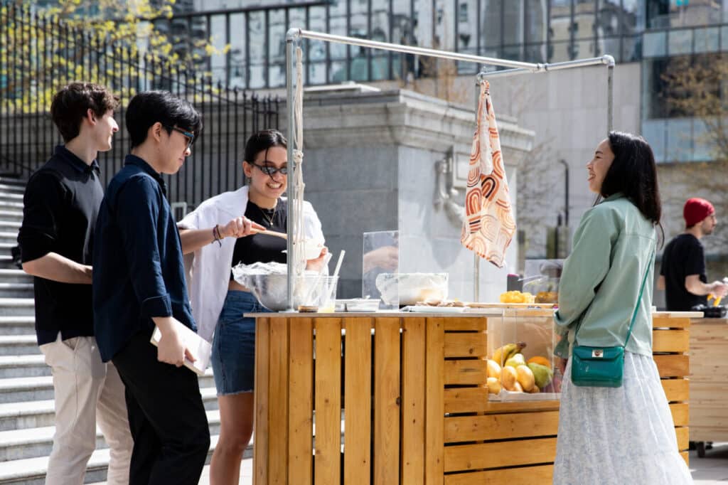 Group of people smiling and serving food at a wooden cart with mangoes displayed underneath, set against the stone steps of a building.