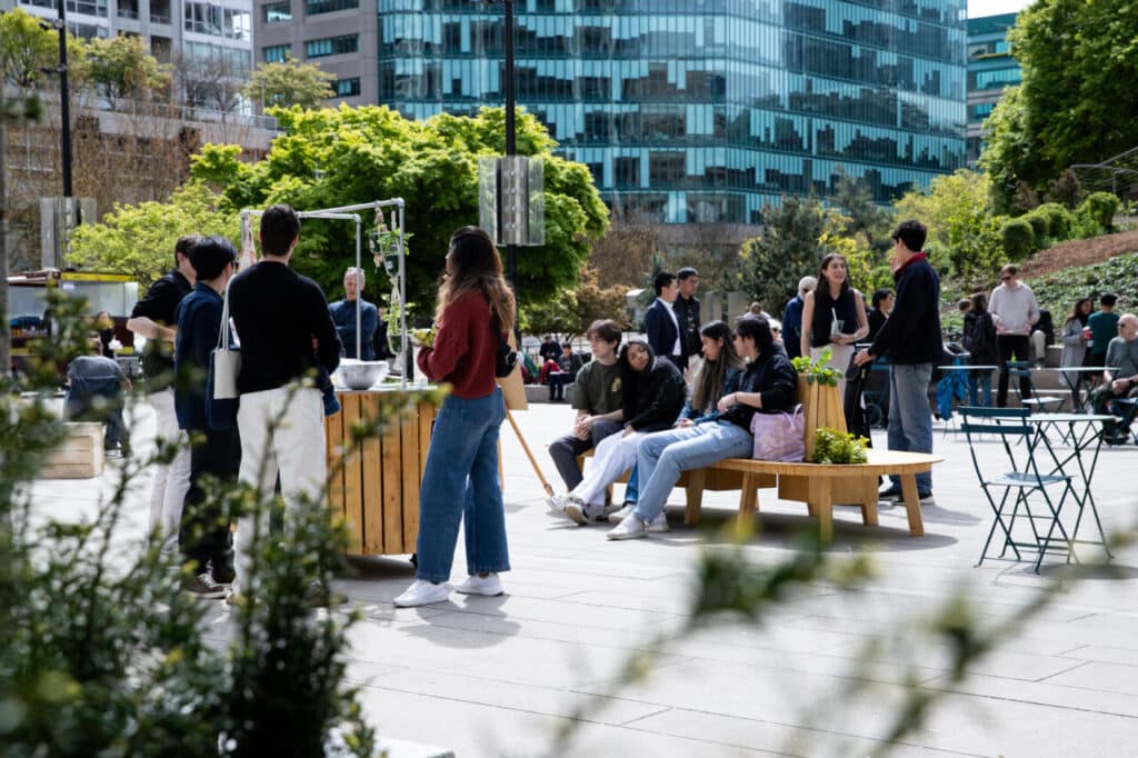Outdoor scene with people gathered around wooden carts and benches in a downtown plaza, surrounded by trees and tall glass buildings.