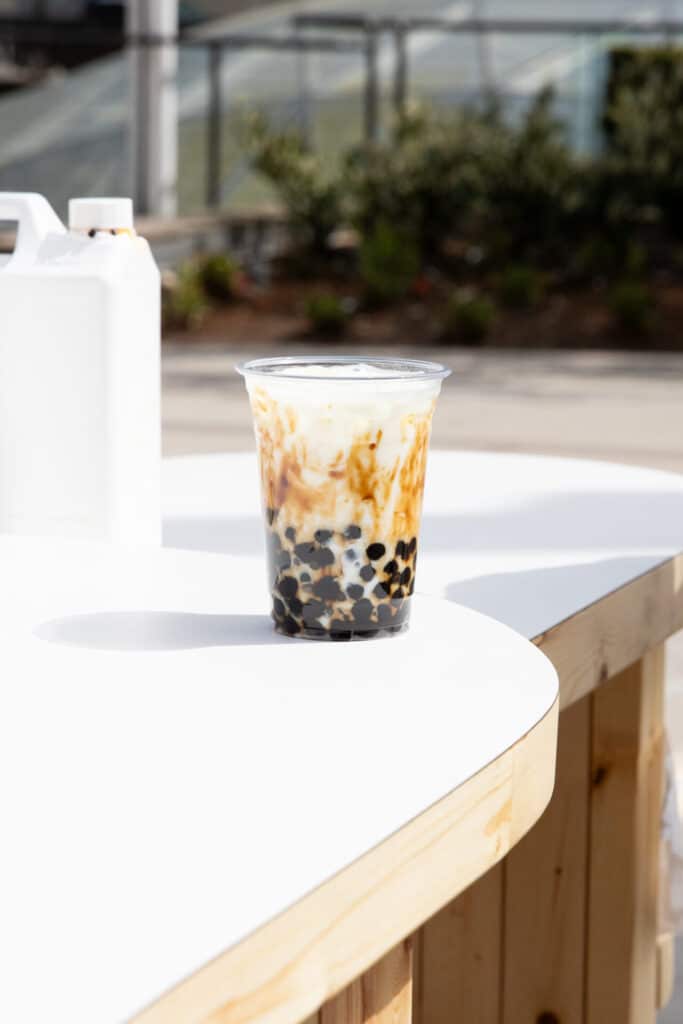 Clear plastic cup of brown sugar bubble tea with tapioca pearls, placed on a white counter in bright sunlight.