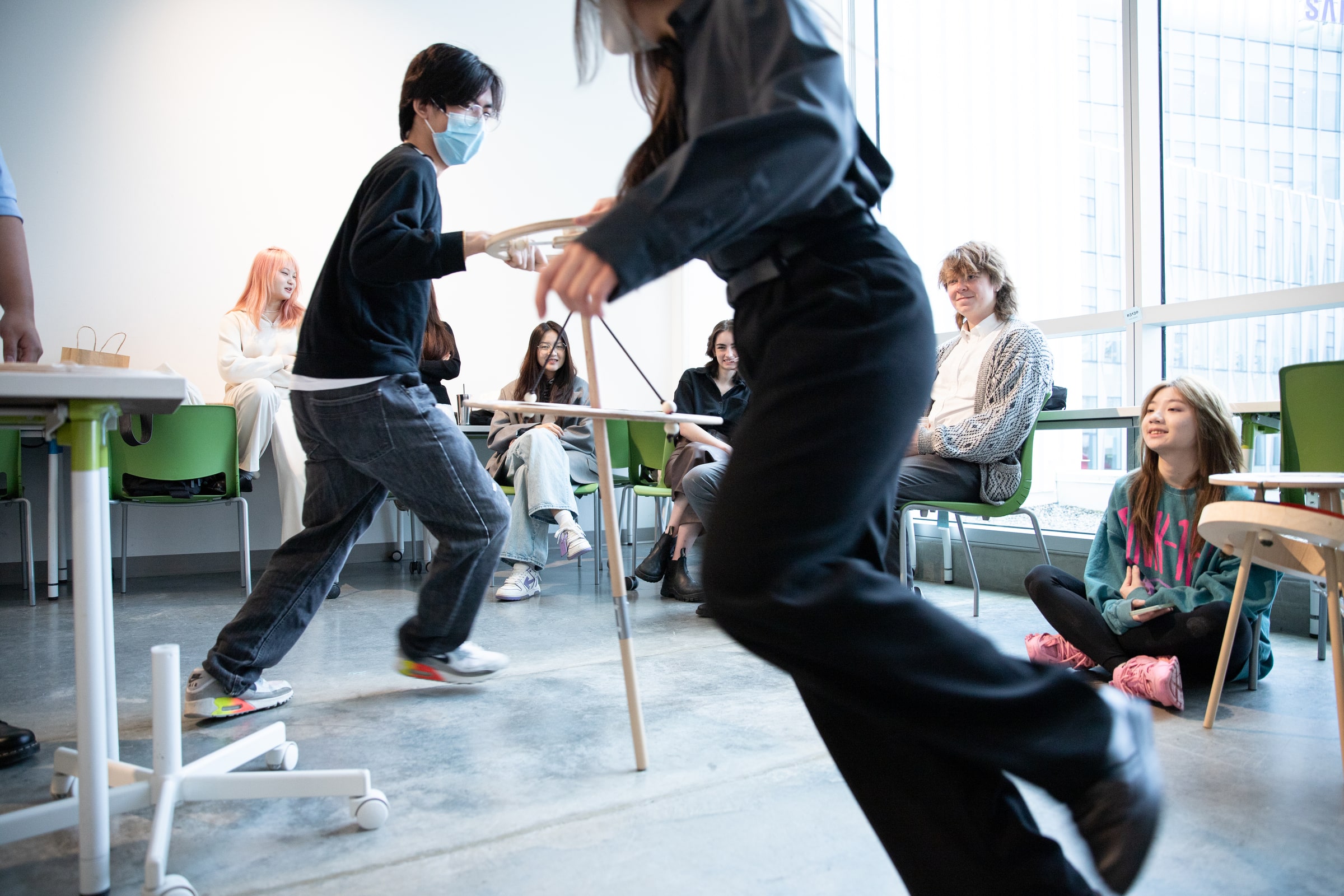 A student in a classroom runs while holding a project prototype as classmates watch and smile during a lively design workshop at Emily Carr University.