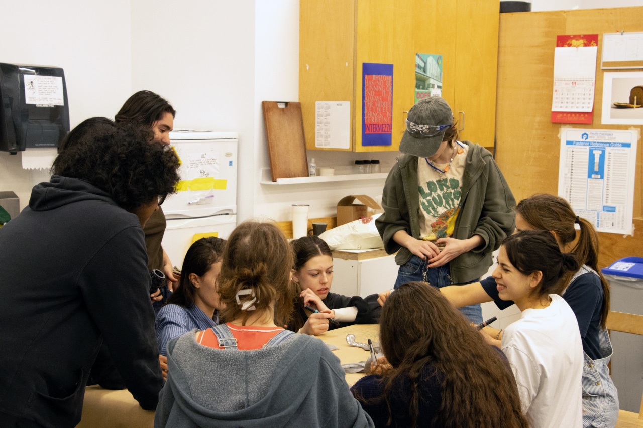 A group of students gather around a worktable, sketching and testing ideas together in a busy design studio.