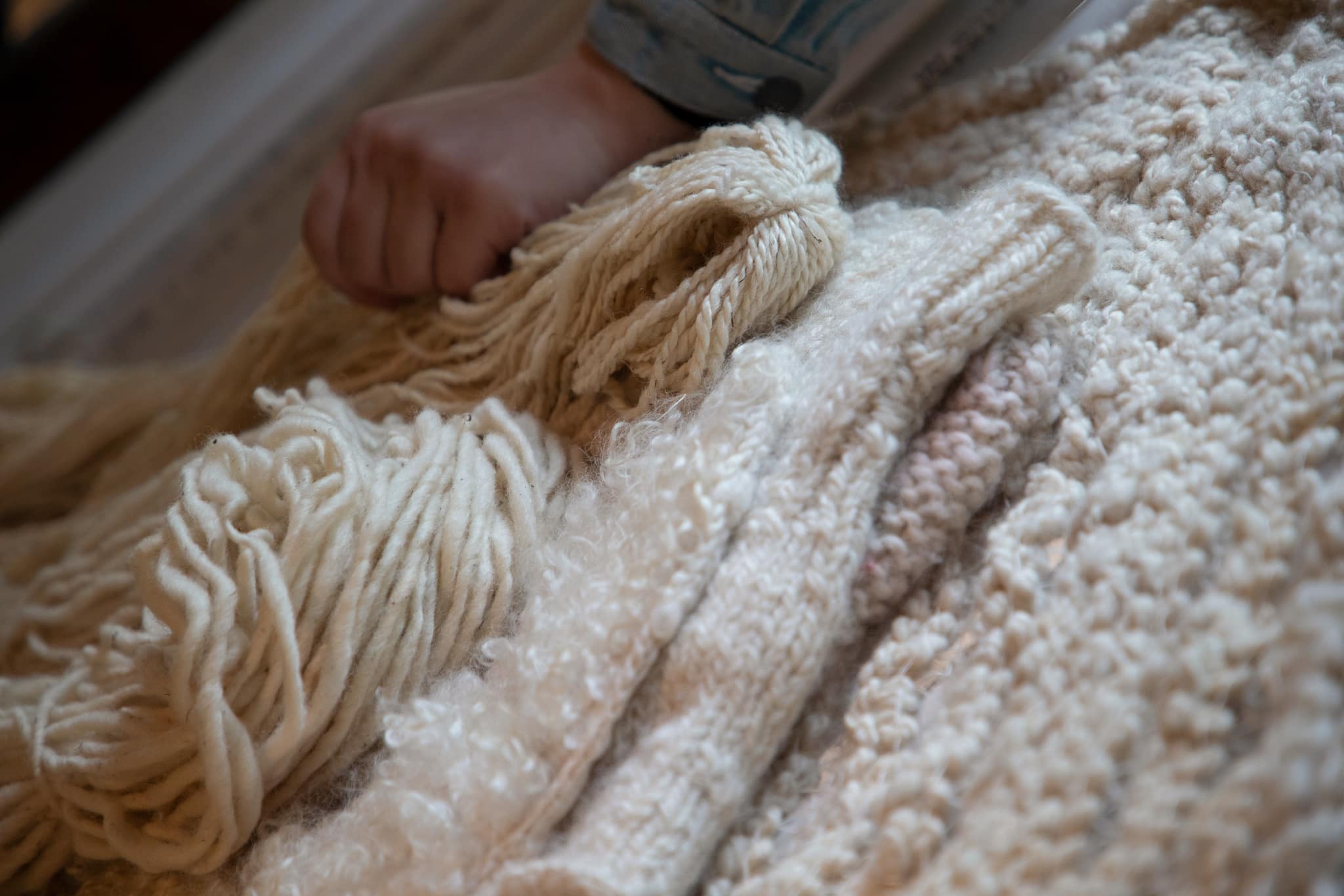 Close-up of a hand gripping thick cream-colored yarn, resting on a textured wool surface with loops and fibers.