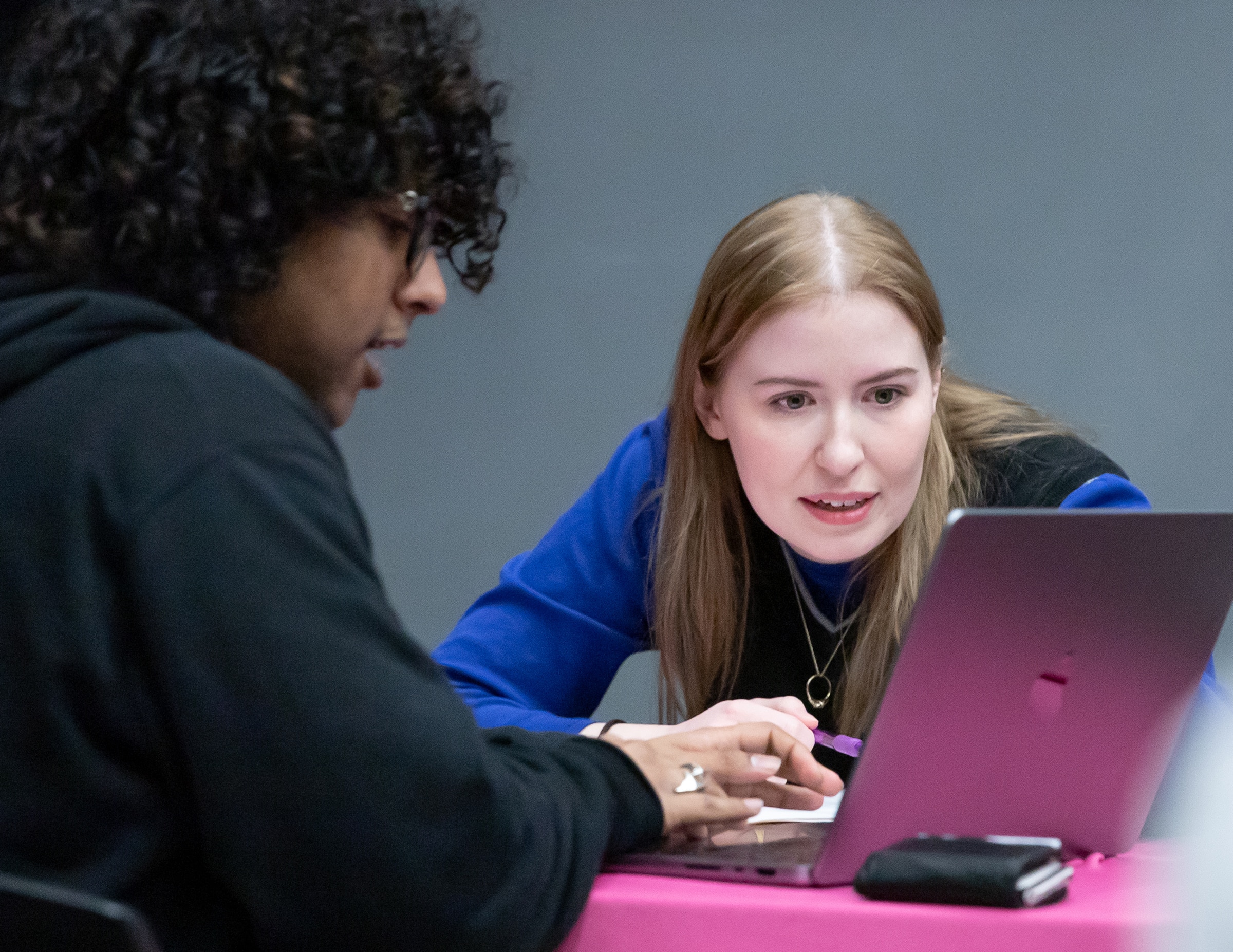 Two students sitting at a table, leaning in toward a laptop. One wears glasses and dark curly hair, while the other, with long blonde hair, points at the screen while explaining something.