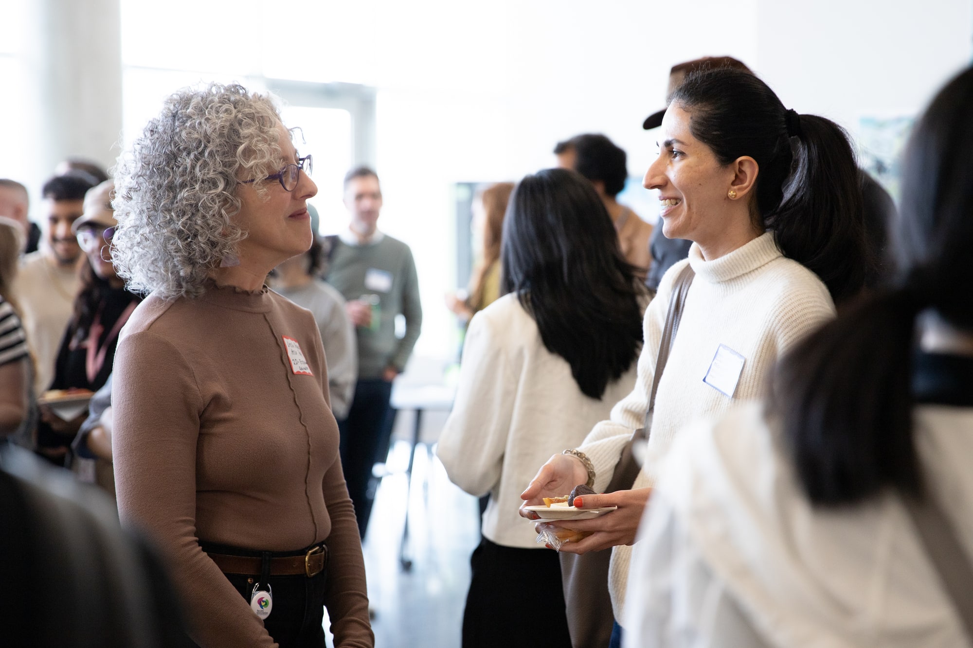 Two women engaged in conversation at a networking event, smiling warmly while surrounded by a crowd of other attendees. One holds a plate of food, and both wear nametags.