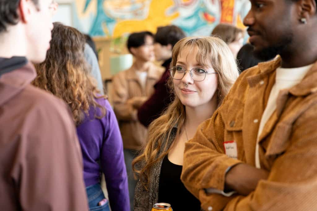 Students talking at a campus gathering; one student with glasses and long hair smiles while listening.