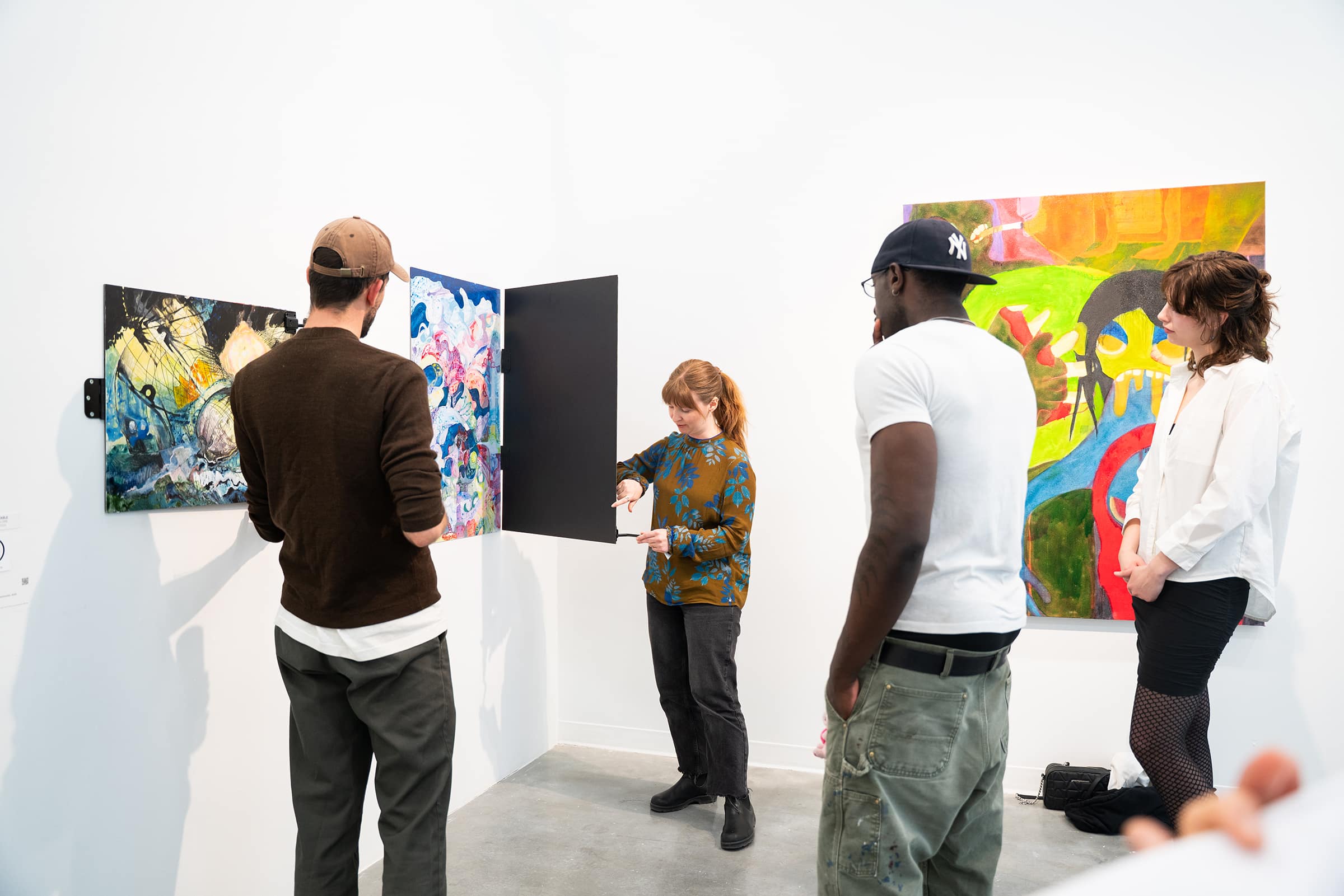 Four people stand in a gallery space observing artwork. A woman in a patterned top demonstrates or adjusts part of a hinged panel artwork that reveals colorful abstract designs. One person in a brown sweater and cap looks closely, while two others, one in a white shirt and cap and another in a white blouse and skirt, watch attentively. A bright, bold painting with abstract figures is visible on the wall behind them.