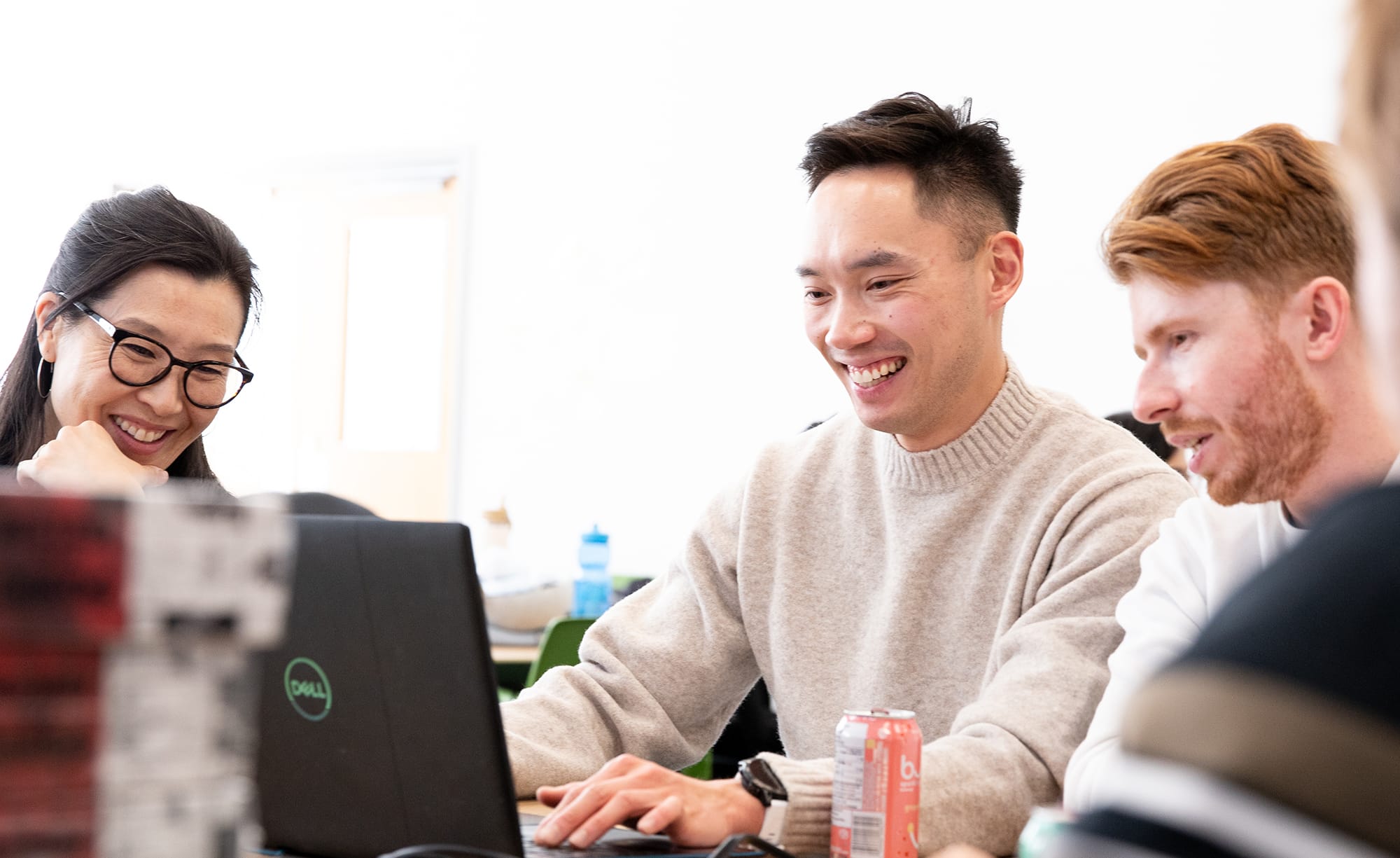 Three people collaborate at a laptop, smiling and engaged in discussion. A soda can and notebooks are on the table.
