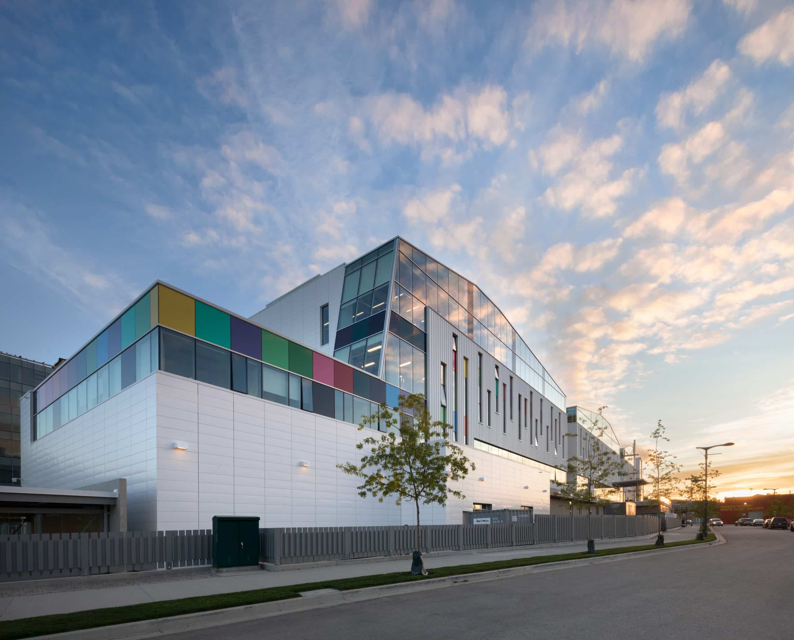 A modern building with colourful panels and large glass windows is shown at sunset, with a partly cloudy sky glowing in the background.