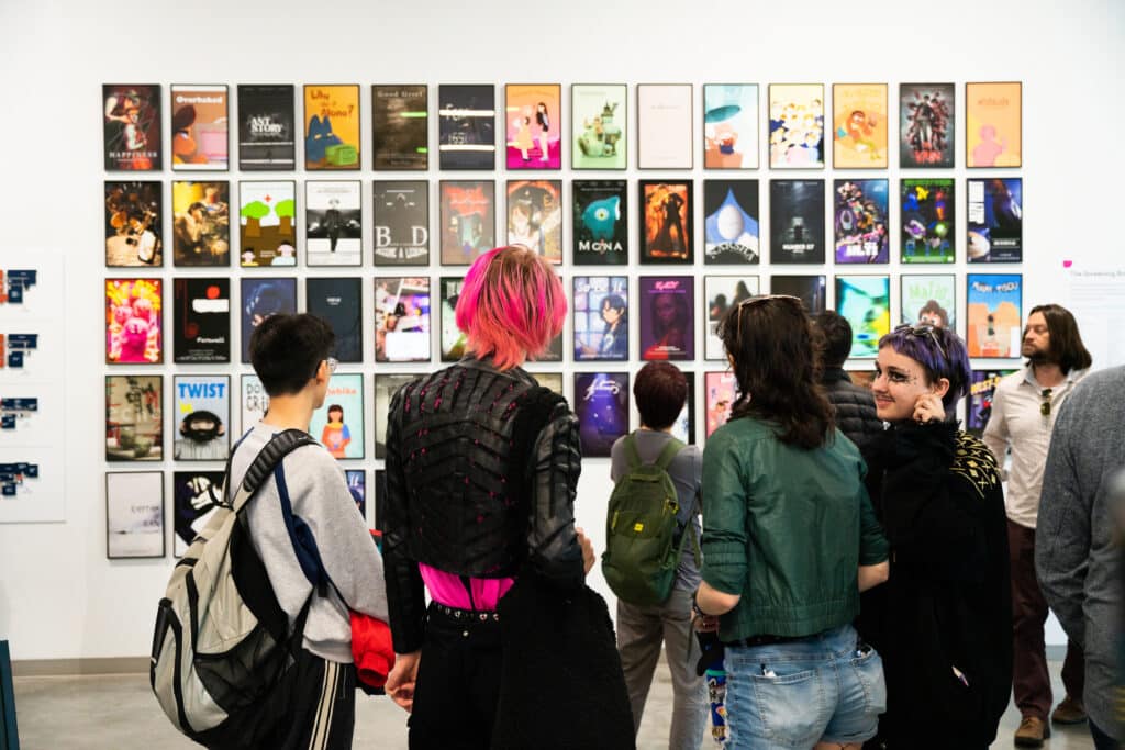 Several people in a gallery setting look at multiple artworks hung on white wall.