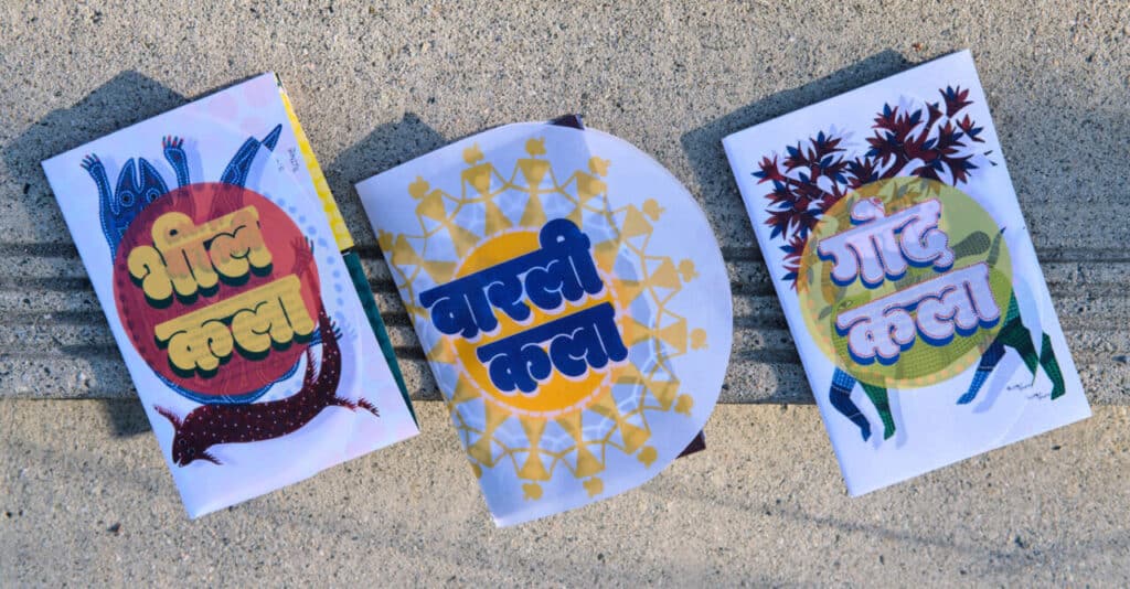 Three illustrated booklets with bold Hindi lettering and traditional patterns displayed side by side on concrete.