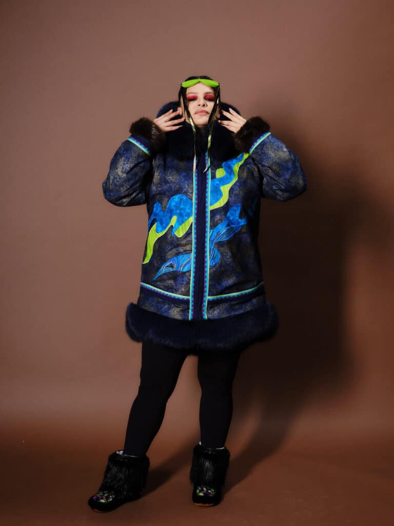 A model poses against a brown backdrop wearing a blue and green coat with fur trim, green goggles, and matching black fur boots.