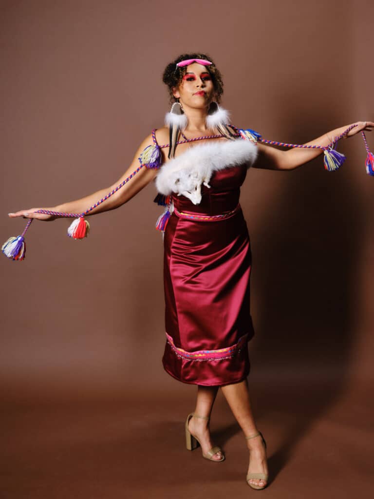 A model in a burgundy satin dress with white fur detail and braided tassels poses with arms outstretched, tassels extended.