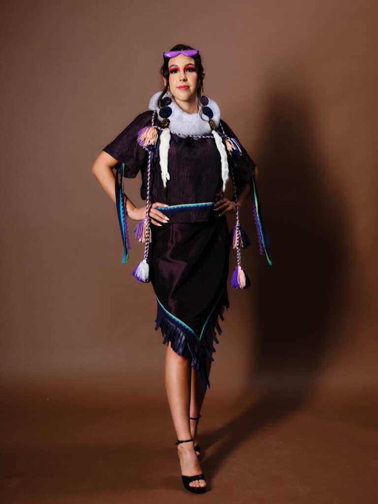 A model in a deep purple dress with fringe and fur details stands with hands on hips, wearing braids and purple accents.