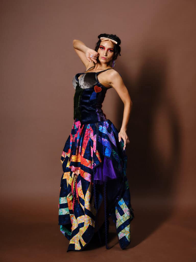 A model poses in a layered multicoloured gown with bright patchwork patterns and tulle, hand raised near face.