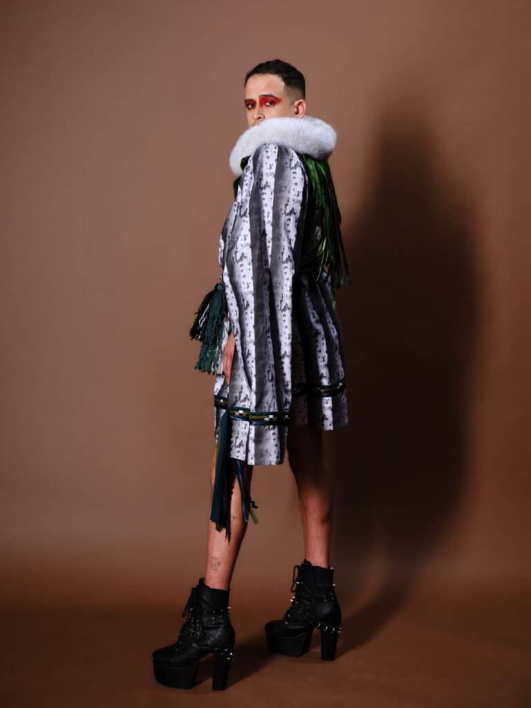 A model in a grey patterned coat with fur collar looks over one shoulder, wearing tall black platform boots.