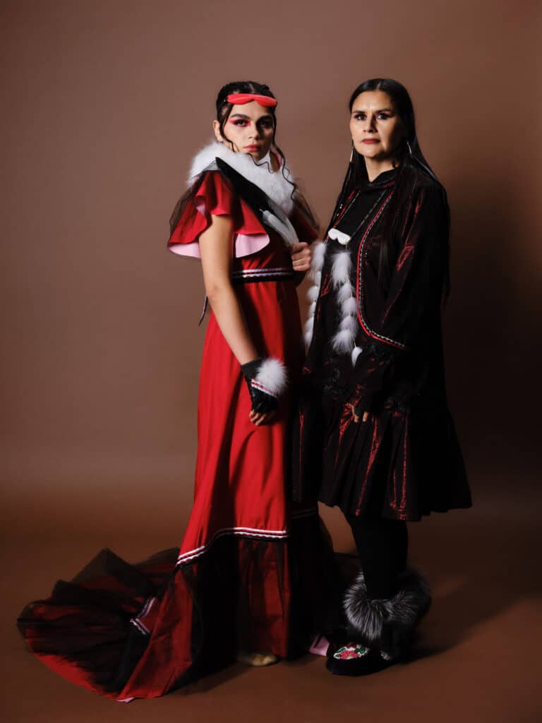 Two models in red and black dresses with fur accents stand side by side against a brown backdrop, their serious expressions emphasizing strength and presence.
