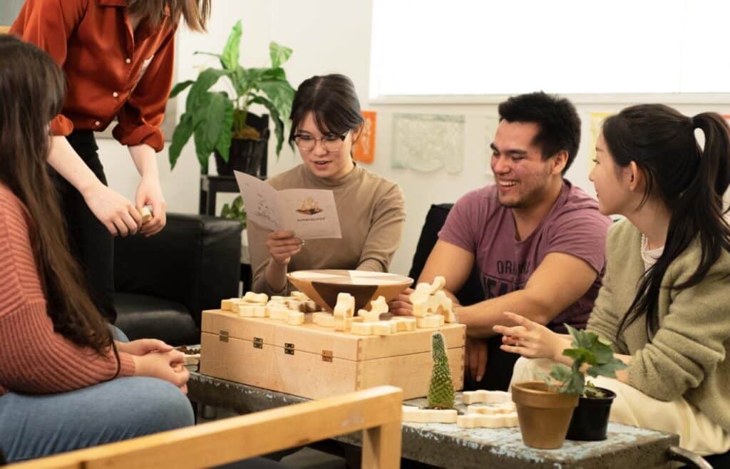 A group of four people gathered around the Genderloop game set, smiling and interacting as they read the instructions and handle the wooden pieces.