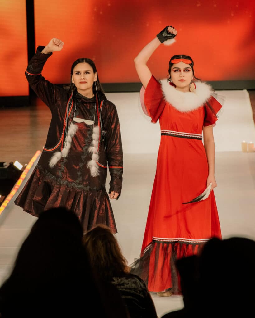 Two models on the runway stand side by side, both raising a fist in solidarity, wearing bold red and dark red dresses with fur and ribbon details.