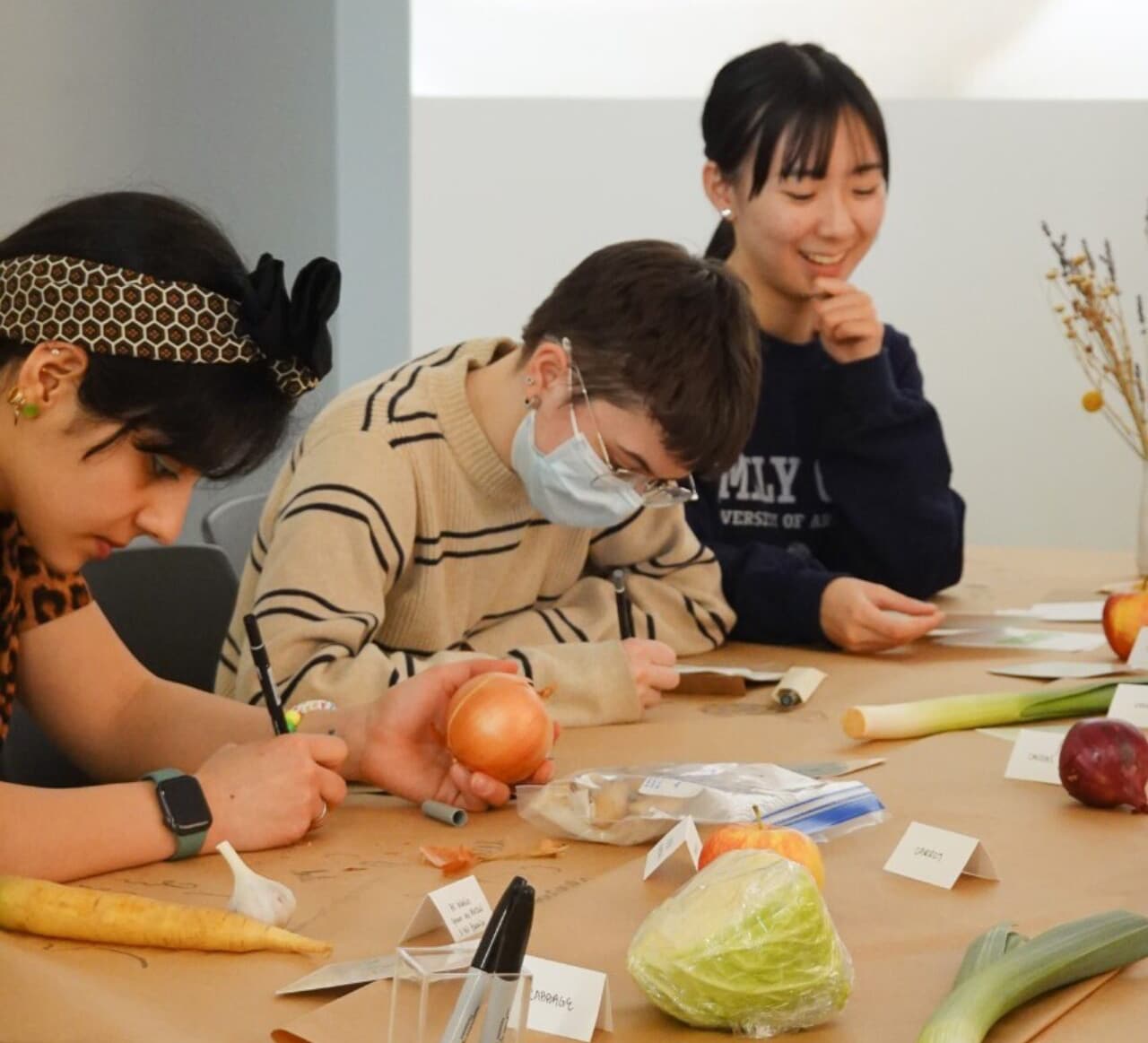 Students sit at a long table covered with vegetables and labels, sketching and writing while experimenting with food-based design.