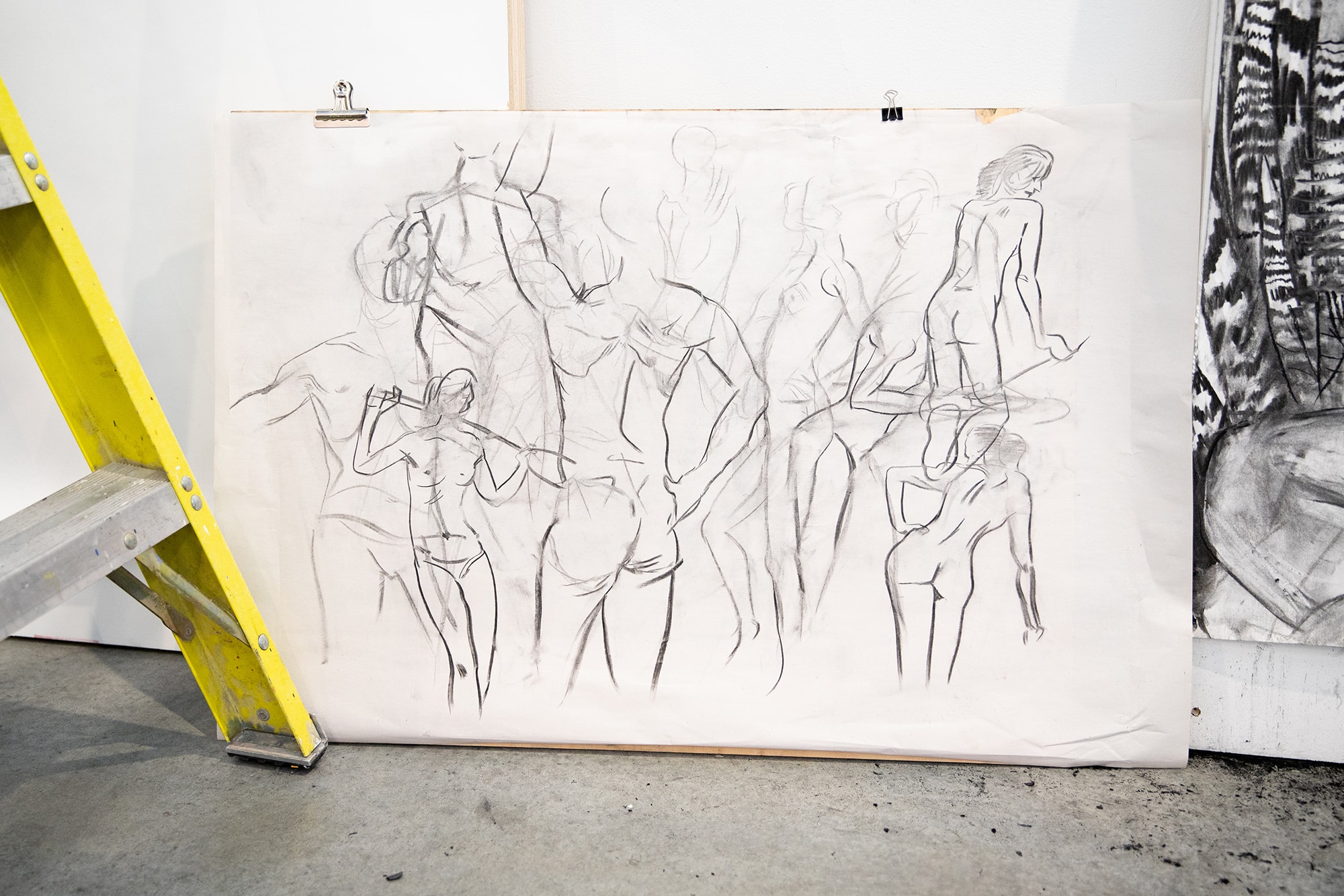 Large sheet of charcoal figure sketches leaning against a wall in a studio, beside a yellow ladder.