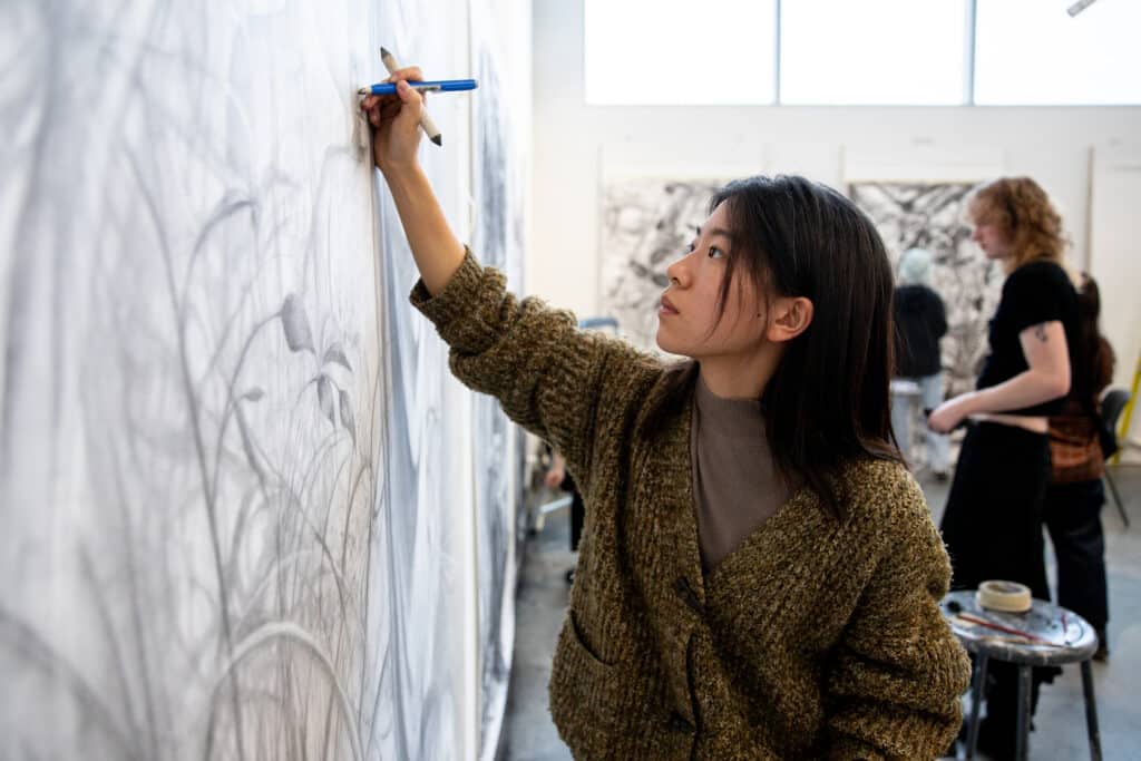 Student drawing with charcoal on a large piece of white paper, backlit by large windows and natural light.