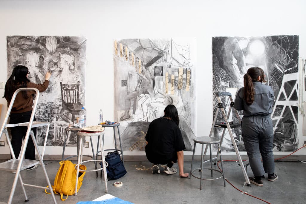 Group of students working on large charcoal drawings taped to the wall, using ladders, stools and drawing tools in a bright studio.