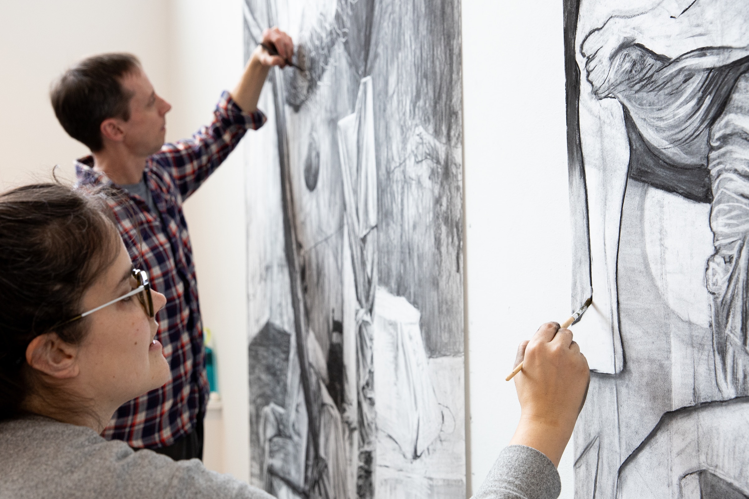 Two people drawing in black and white on oversized pieces of white paper.
