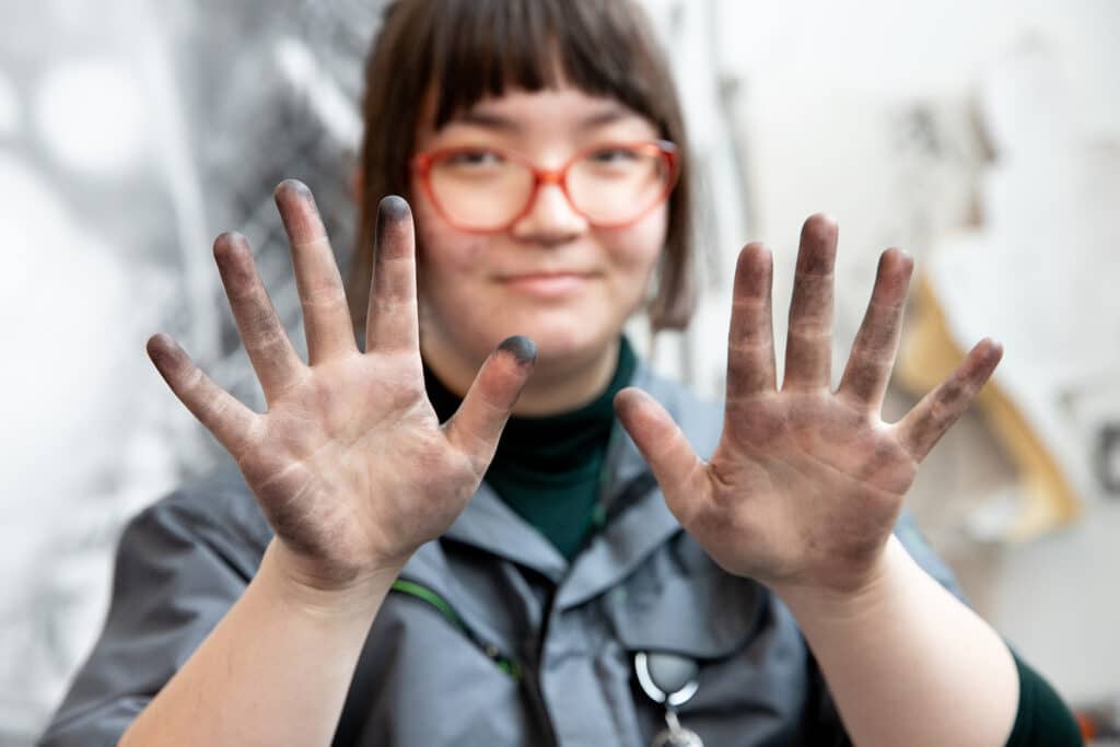 Close-up of a student with red glasses holding up both hands, palms out, showing fingers smudged with dark charcoal.