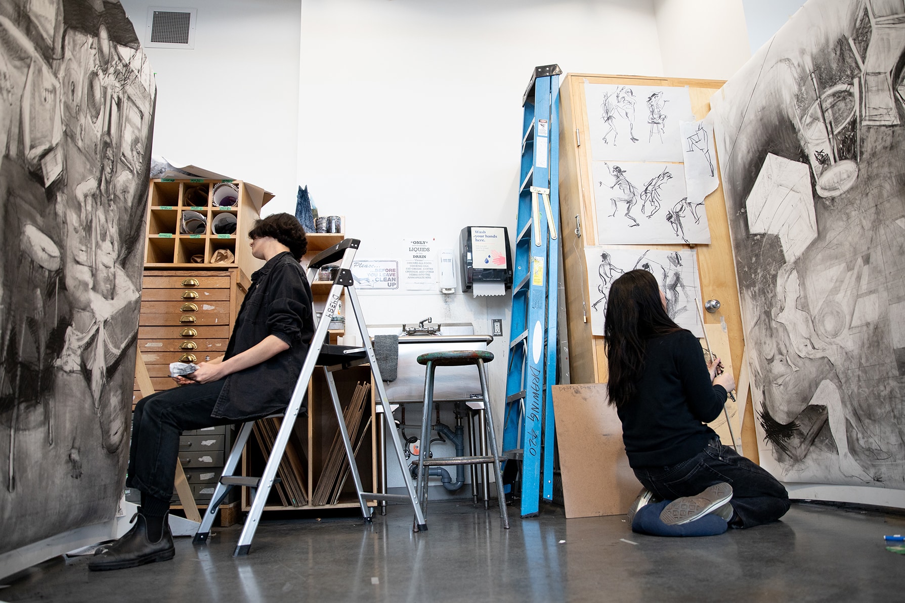 Students seated and kneeling in a studio, working on large charcoal drawings that cover tall boards, with figure sketches pinned to the wall.